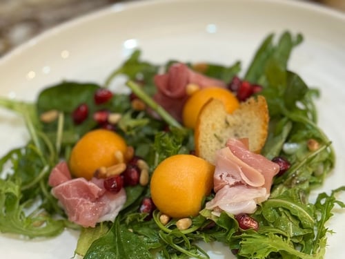 Fresh salad with prosciutto, melon, pomegranate seeds, pine nuts, and croutons.