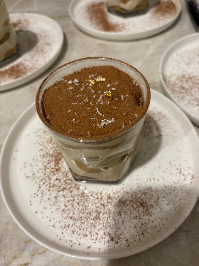 Individual tiramisu dessert cup with cocoa powder and gold flakes.